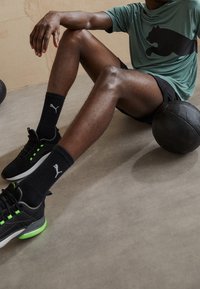 Czarne buty sportowe z zielonymi akcentami, czarne skarpetki, czarne szorty oraz turkusowa koszulka z dużym czarnym logo kota, siedzący z piłką do koszykówki.