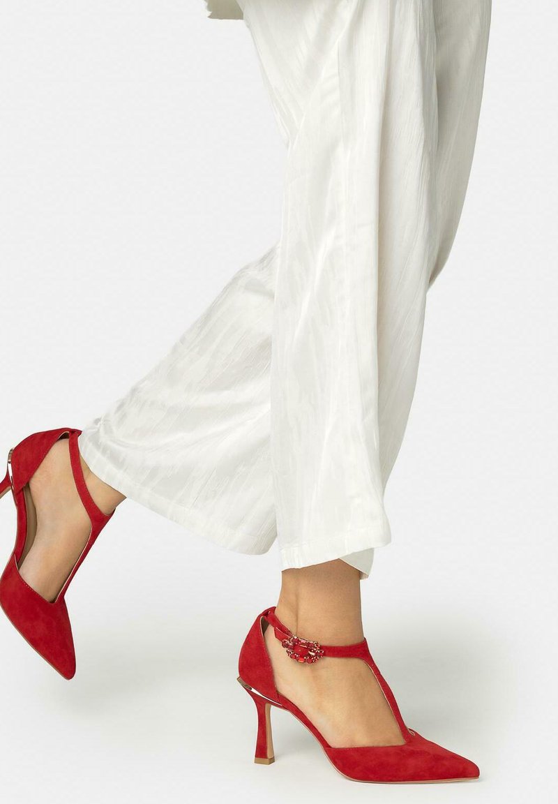 Personne portant un pantalon blanc à jambes larges et des chaussures à talons hauts rouges en daim avec bride en T ornementée d'une boucle, un pied légèrement levé.
