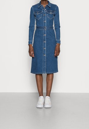 Kobieta w niebieskiej sukience dżinsowej do połowy łydki z długimi rękawami, z przodu zapinanej na guziki, z kieszeniami na piersi, paskiem i białymi trampkami.
