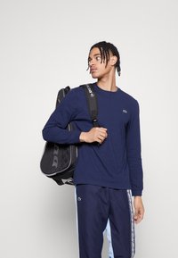 Μπλούζα ναυτικού στιλ με μακρύ μανίκι και στρογγυλή λαιμόκοψη, κεντημένο λογότυπο, σε συνδυασμό με σκούρες μπλέ και ανοιχτό γαλάζιες φόρμες με πλαϊνές ρίγες, και μαύρη αθλητική τσάντα.