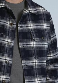 Marineblauw en witte geruite flanellen shirt met knoopsluiting, borstzakken en een grijze ronde hals eronder. Gestructureerde stof met zichtbare stiksels.
