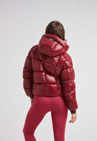 Giacca piumino rossa con finitura lucida, design trapuntato strutturato, cappuccio staccabile e polsini elastici. Indossata con leggings rosa abbinati.