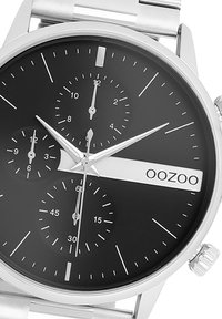 Montre en acier inoxydable argenté avec un cadran noir, présentant trois sous-cadrans, des index horaires argentés et un logo "OOZOO" blanc sur le côté.