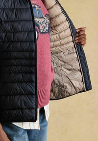 Melnā pārklātā jaka ar beidzējo iekšpusi. zem tās ir kontrastējošs ribots rozā džemperis. Iekšpusē ir redzami etiķetes.