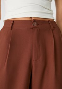 Bruine op maat gemaakte broek van gladde stof, met een sluiting aan de voorkant met een knoop, zijzakken en scherpe plooi-details.