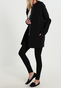 Černý dlouhý kabát s předním zipem, kulatým límcem a dlouhými rukávy, doplněný o černé úzké džíny a ploché boty.