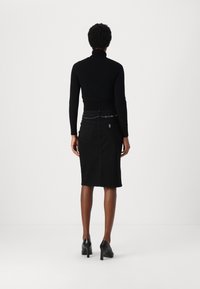 Mujer vestida con un suéter negro de cuello alto, una falda negra hasta la rodilla con un cinturón de cadena y zapatos de tacón altos negros, de pie mirando hacia atrás.