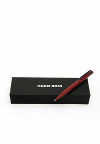 Roter, eleganter Stift mit schwarzem Clip, der auf einer schwarzen Box liegt, die mit der weißen Marke "HUGO BOSS" versehen ist. Die Box ist rechteckig und minimalistisch.