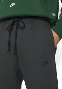 Graue Nike-Sweatpants mit glatter Textur, ausgestattet mit einem schwarzen Logo und einem verstellbaren Kordelzug. Hand eines Mannes, die sich auf die Tasche stützt.
