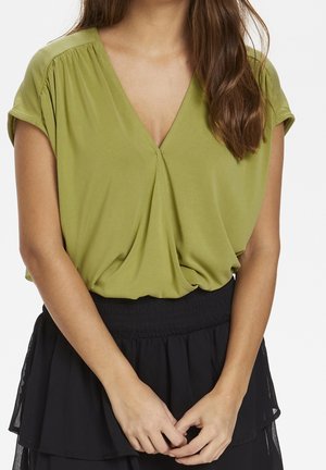 Vrouw draagt een losse olijfgroene V-hals blouse met opgerolde mouwen, gestoken in een zwarte gelaagde rok met hoge taille, handen voor zich samengevouwen.