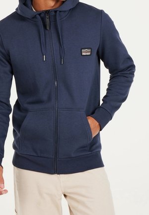 Uomo che indossa una felpa con cappuccio blu navy con zip e tasche frontali, con un piccolo logo "Protest" sul petto, abbinata a pantaloni beige chiaro.