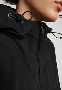 Schwarze Kapuzenjacke mit glatter Textur, Reißverschluss, verstellbarem Kordelzug und dezenten Nahtdetails.