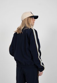 Femme aux cheveux blonds portant une casquette beige et marine ainsi qu'un survêtement marine avec des rayures blanches et jaunes, vue de profil sur un fond uni.