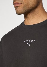 Juodas medvilnės marškinėliai su apvaliu kaklu, ant kurių baltu siuvinėjimu užrašyta "HYROX", o žemiau teksto yra mažas Puma logotipas.