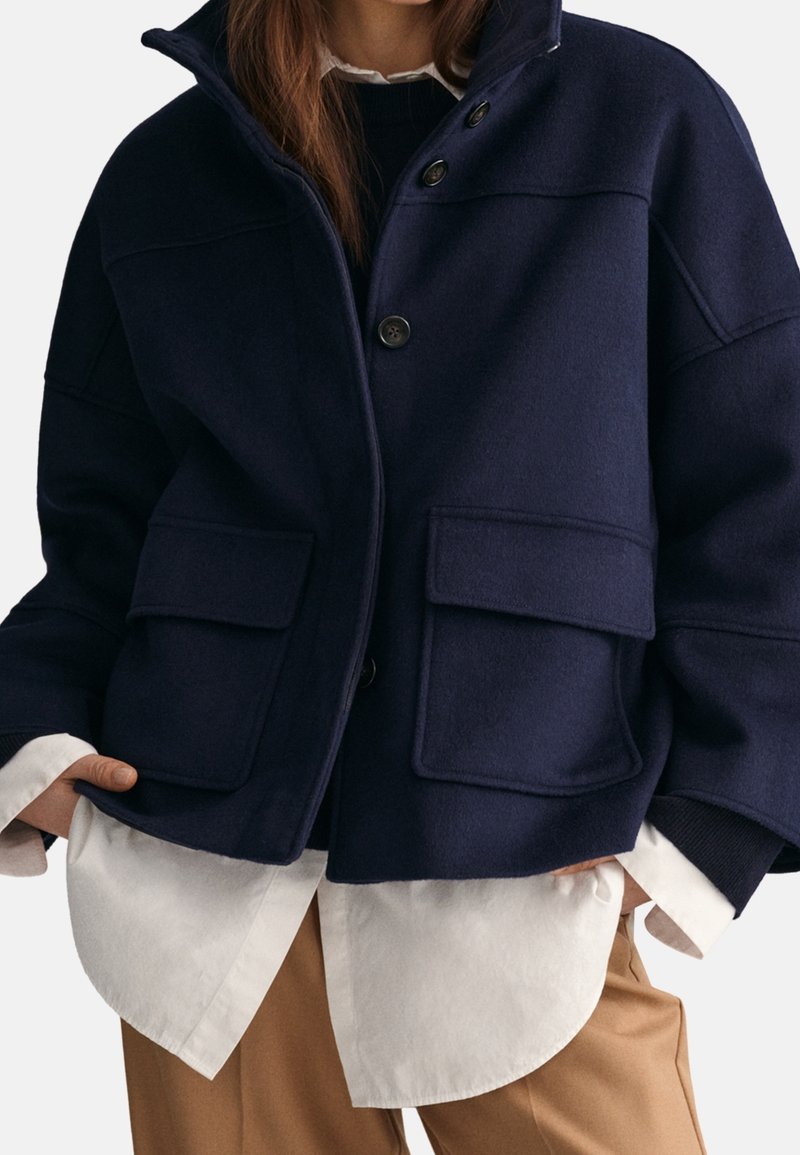 Giacca in misto lana blu navy con colletto alto, chiusura a bottoni e due tasche applicate frontali. Indossata sopra una camicia bianca e pantaloni beige.