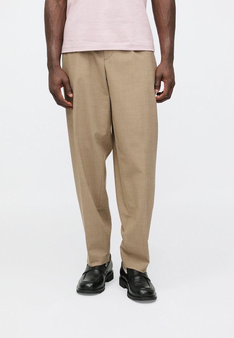 Calças bege feitas à medida com design de perna reta, fabricadas em tecido leve. Combinadas com sapatos de couro pretos e uma t-shirt cor-de-rosa clara.