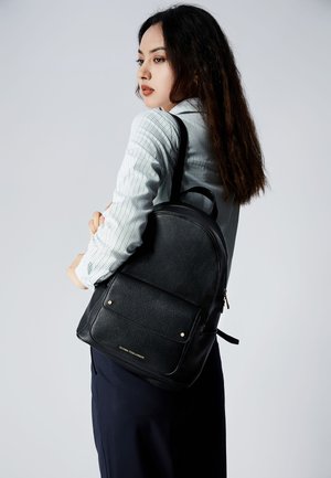 Zaino in pelle nera con texture a ciottoli, forma rotonda, tasca frontale con hardware dorato, branding minimale, indossato sulla spalla.