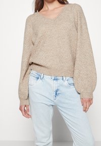 Frau trägt einen beigen, strukturierten V-Ausschnitt-Pullover mit langen Ballonärmeln und hellblauen, hochtaillierten Jeans.