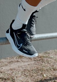 Fekete és szürke sportcipők szőtt textúrával, fehér részletekkel és fűzőkkel, vízszintes rúdra helyezve, fehér zoknikkal párosítva.