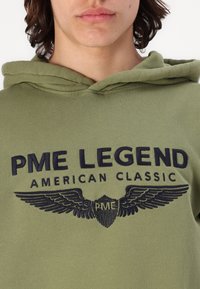 Zielona bluza z kapturem z haftem z przodu z napisem "PME LEGEND" oraz "AMERICAN CLASSIC", ozdobiona grafikami skrzydeł w granatowym kolorze. Miękka tekstura materiału.