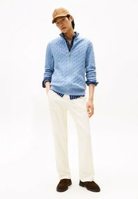 Helles blauer Kabelstrickpullover mit Reißverschluss, über einem marineblauen gestreiften Hemd getragen, kombiniert mit weißen Hosen und braunen Slippern. Braune Mütze auf dem Kopf.