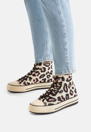 Osoba nosząca jasnoniebieskie dżinsy i wysokie trampki z czarno-różowym wzorem lamparta na beżowym tle.