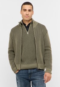 Kakhi-grön stickad tröja med zip-up krage, med vertikal ribbad textur och avslappnad passform. Bärs över en svart rutig skjorta.