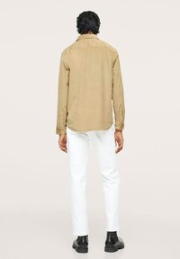 Mand stående med ryggen til, iført en beige langærmet skjorte, hvide bukser og sorte ankelstøvler foran en ensfarvet baggrund.