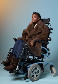 Μια αυτοπεποίθηση γυναίκα με καφέ γούνινο παλτό και μπότες καθισμένη σε ένα μπλε ηλεκτρικό καροτσάκι πίσω από ένα φόντο σε κλίμακα μπλε και πορτοκαλί.