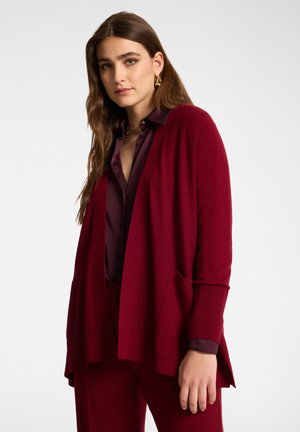 Frau mit langen braunen Haaren, die ein geschichtetes dunkelrotes Outfit trägt, bestehend aus einem Cardigan, Hemd und Hose, vor einem einfarbigen Hintergrund stehend.