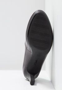 Scarpa con tacco alto in pelle marrone con punta affusolata, caratterizzata da una suola nera testurizzata e sottili dettagli di cucitura. Posata su una superficie bianca.