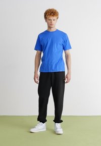 Camiseta de algodón azul con manga corta y cuello redondo, combinada con pantalones de chándal negros. El modelo lleva zapatillas deportivas blancas y está de pie sobre un suelo verde.
