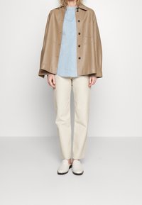 Ljusbrun läderkappa över en ljusblå skjorta med knappar, i kombination med krämfärgade byxor och vita slip-on skor.