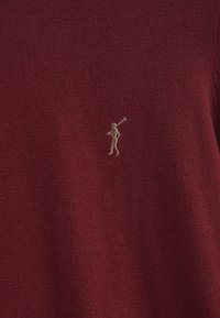 Burgunderfarbener Strickpullover mit glatter Textur und kleinem beigefarbenem Logo einer Figur, die einen Golfschläger hält, auf der linken Seite.