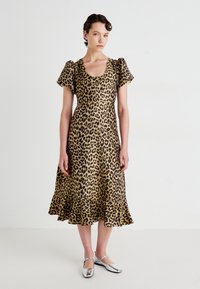Žena stojí oblečená v midi šatách s leopardím vzorom a balónovými rukávmi, v strieborných Mary Jane topánkach na pozadí bielej farby.