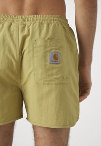 Pantalones cortos de cinturilla elástica en tela amarilla clara, con un bolsillo trasero que tiene un parche de logo de Carhartt en gris y naranja y dos detalles de botón metálico.