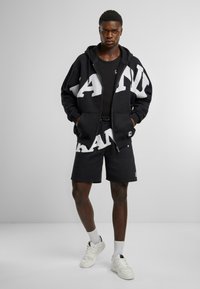 Black zip-up hoodie with large white lettering, paired with matching black shorts. Features a relaxed fit and front pockets. White sneakers and socks.