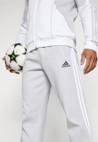 Tuta grigia chiara con strisce bianche Adidas, giacca con zip e pantaloni con tasche laterali. Una palla da calcio è tenuta in mano con un design a stella nera.