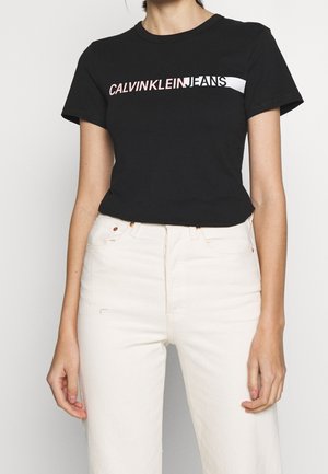 Woman wearing a black Calvin Klein Jeans t-shirt and high-waisted white jeans, standing against a plain background.