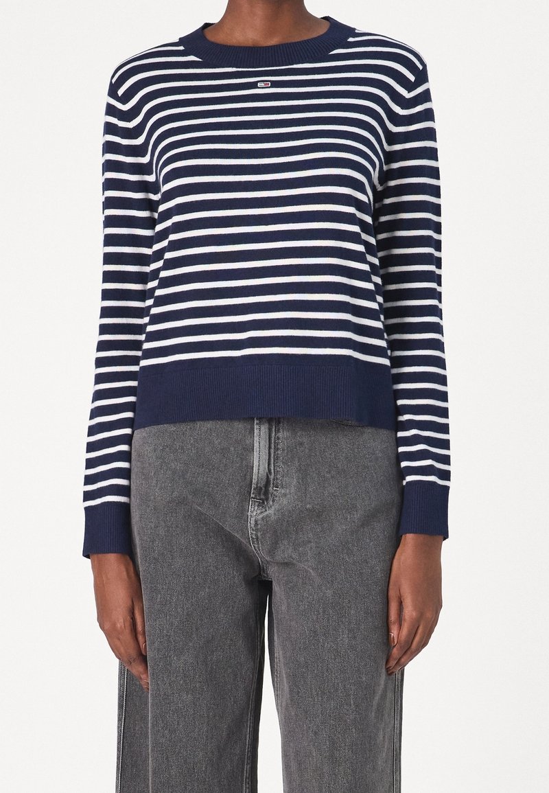 Osoba ubrana w granatową i białą w paski bluzę z długim rękawem, z granatowymi mankietami i dołem, zestawioną z wysokimi, wyblakłymi czarnymi dżinsami.