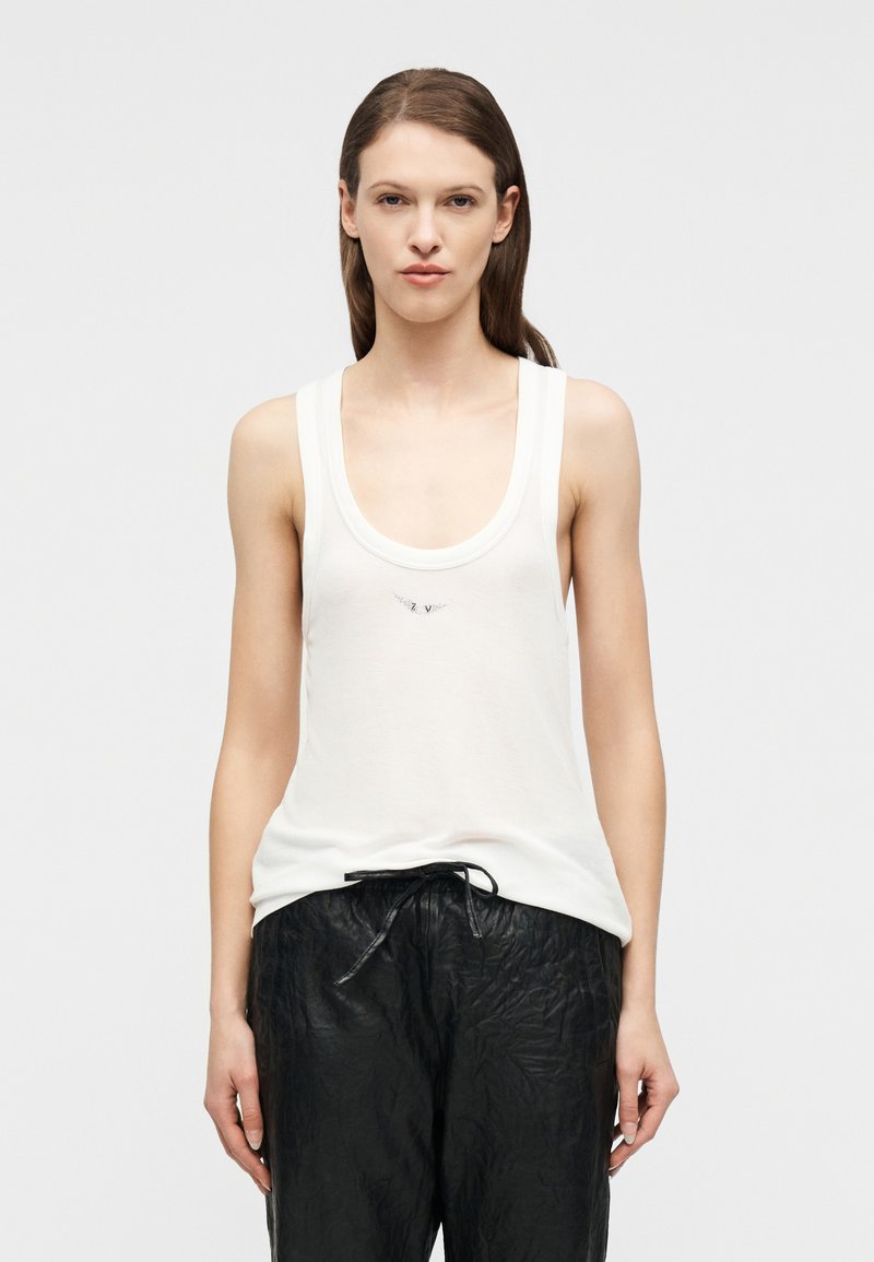 Γυναίκα με ίσια καστανά μαλλιά φοράει λευκό αμάνικο τοπάκι και φαρδιά μαύρη παντελόνα με κορδόνι, στέκεται μπροστά σε απλό φωτεινό φόντο.