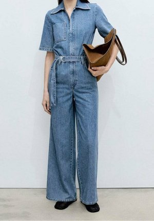 Jeansowy kombinezon z krótkimi rękawami, kieszenią na piersi i paskiem w talii. Jasnoniebieski kolor, szerokie nogawki, w połączeniu z brązową torbą na ramię.