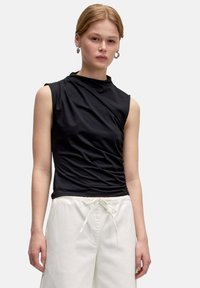 Zwarte mouwloze top met een hoge hals, voorzien van geplooide details en een slim fit, gecombineerd met losse witte broeken.