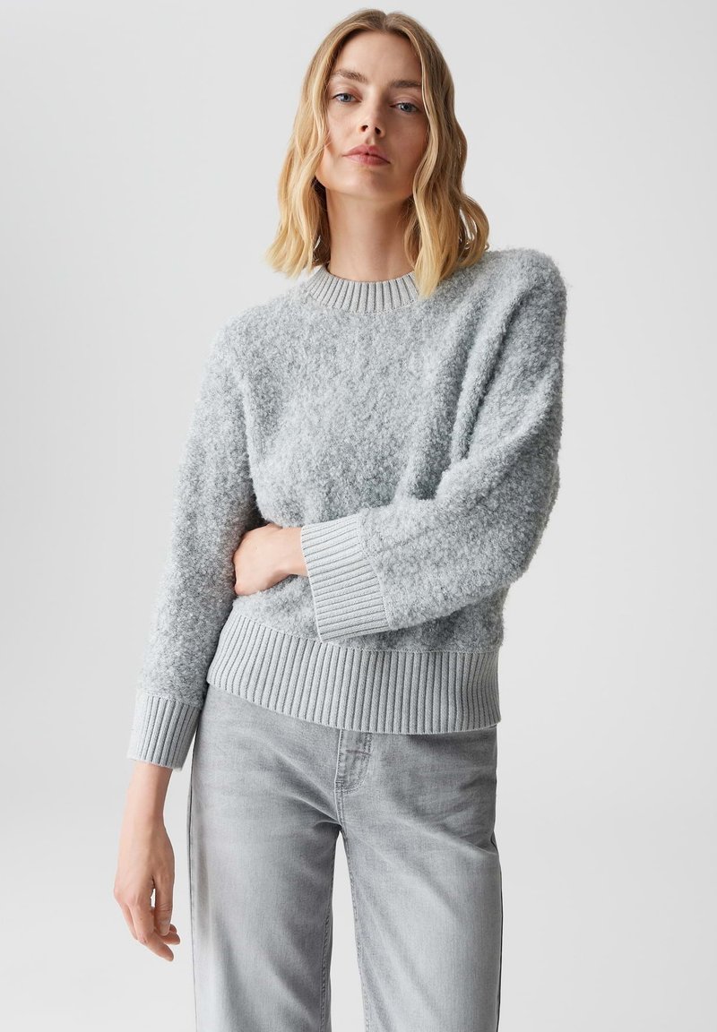 Helles graues Sweatshirt aus einem strukturierten, flauschigen Material. Ausgestattet mit einem gerippten Halsausschnitt und gerippten Bündchen sowie einem gerippten Saum. Kombiniert mit hellgrauen Jeans.