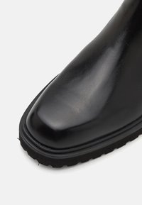 Czarne skórzane buty z gładką, błyszczącą cholewką, zaokrąglonym noskiem oraz grubą, teksturowaną czarną gumową podeszwą. Bez widocznych elementów metalowych ani akcentów.