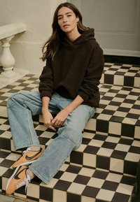 Brązowa polarowa bluza z kapturem, luźne jasnoniebieskie dżinsy i pomarańczowe sneakersy Nike. Siedzi na czarno-białych w kratkę stopniach. Styl casual.