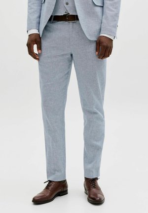 Mann iført lysegrå dressbukser, brun skinnbelte og polerte brune dressko, stående mot en ensfarget hvit bakgrunn.