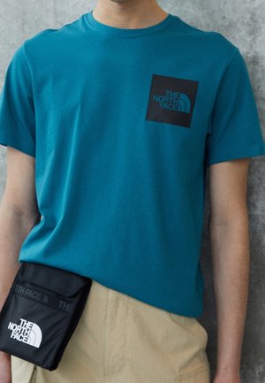 Person iført en turkis The North Face t-shirt og beige bukser med sort The North Face crossbody taske mod en grå baggrund.
