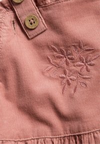 Rosa kordväv med blommönster i en mörkare nyans. Två ljusbruna knappar är synliga upptill, med en volangkant.