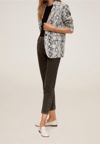 Blazer imprimé serpent en blanc, gris et noir, associé à un jean foncé. La tenue est complétée par des chaussures blanches sans lacets.